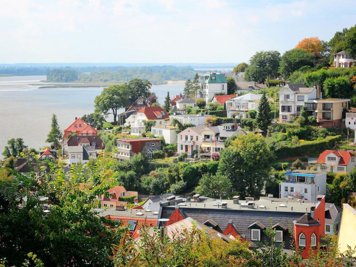 Landscape view of Blankenese with colourful houses, lush greenery and a river in the background under a partly cloudy sky.