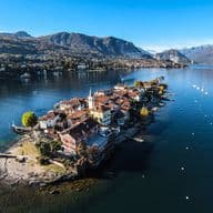 Aerial view of Isola dei Pescatori (Fishermen's Island), a small inhabited island with traditional buildings and red-tiled roofs extending into Lake Maggiore. The island features a church with a bell tower and is surrounded by clear blue water with a few boats. In the background are dramatic mountains and the shoreline town, creating a picturesque Italian lake landscape under a bright blue sky.