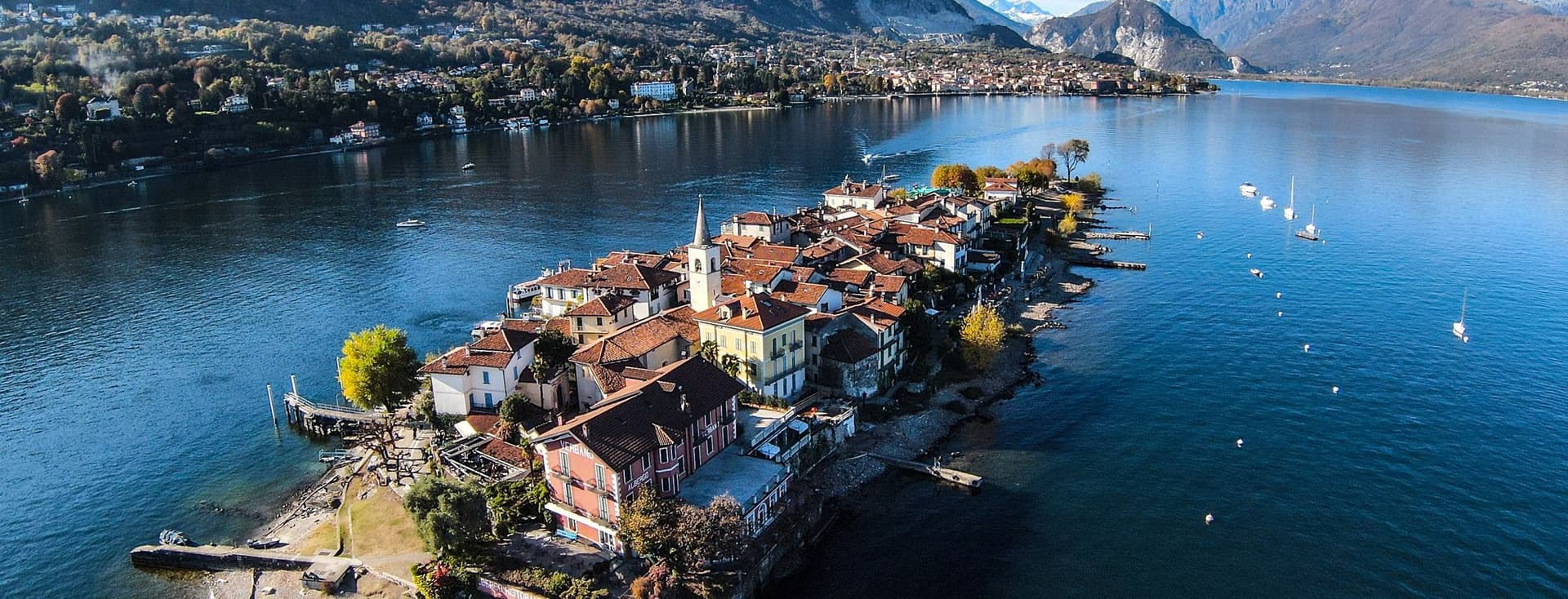 Aerial view of Isola dei Pescatori (Fishermen's Island), a small inhabited island with traditional buildings and red-tiled roofs extending into Lake Maggiore. The island features a church with a bell tower and is surrounded by clear blue water with a few boats. In the background are dramatic mountains and the shoreline town, creating a picturesque Italian lake landscape under a bright blue sky.
