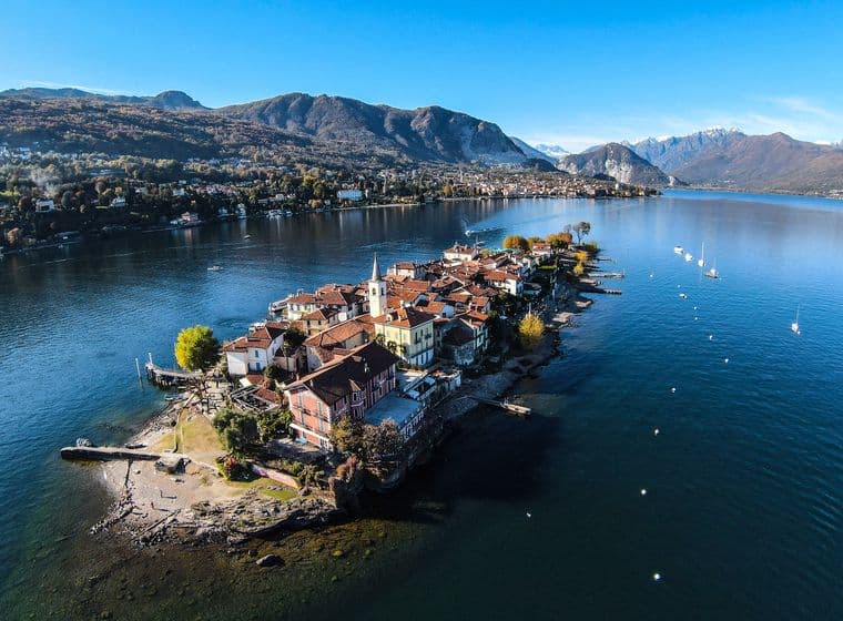 Luftaufnahme der Isola dei Pescatori (Fischerinsel), einer kleinen bewohnten Insel mit traditionellen Gebäuden und roten Ziegeldächern, die in den Lago Maggiore hineinragt. Die Insel zeigt eine Kirche mit Glockenturm und ist umgeben von klarem blauen Wasser mit einigen Booten. Im Hintergrund befinden sich dramatische Berge und eine Küstenstadt, die eine malerische italienische Seelandschaft unter einem strahlend blauen Himmel bilden.