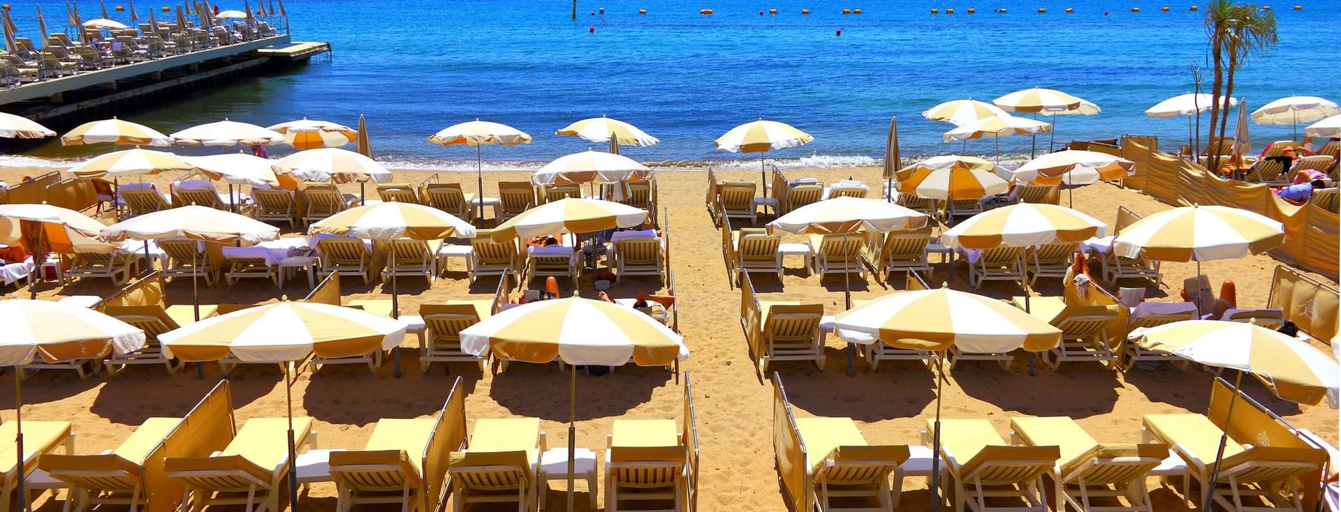 Beach scene with rows of yellow and white umbrellas and lounge chairs on sand. Blue sea with cruise ship and lighthouse in background.