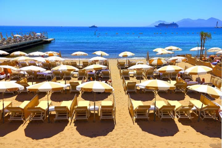 Scène de plage avec des rangées de parasols jaunes et blancs et des chaises longues sur le sable. Mer bleue avec un bateau de croisière et un phare en arrière-plan.