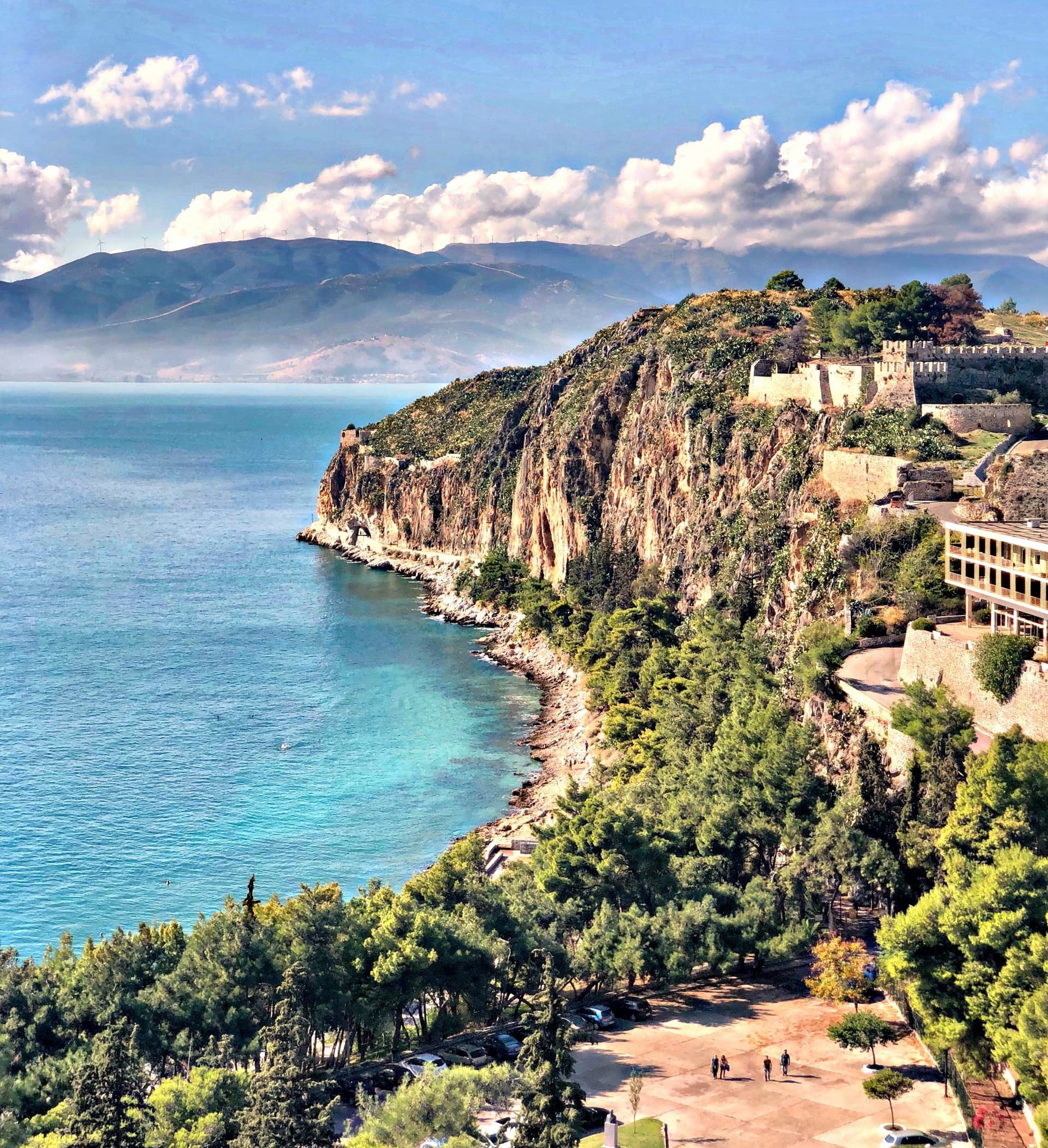 Scenic view of Nafplio, Greece, featuring a rocky coastline, turquoise sea, and a fortress atop a hill under a partly cloudy sky.