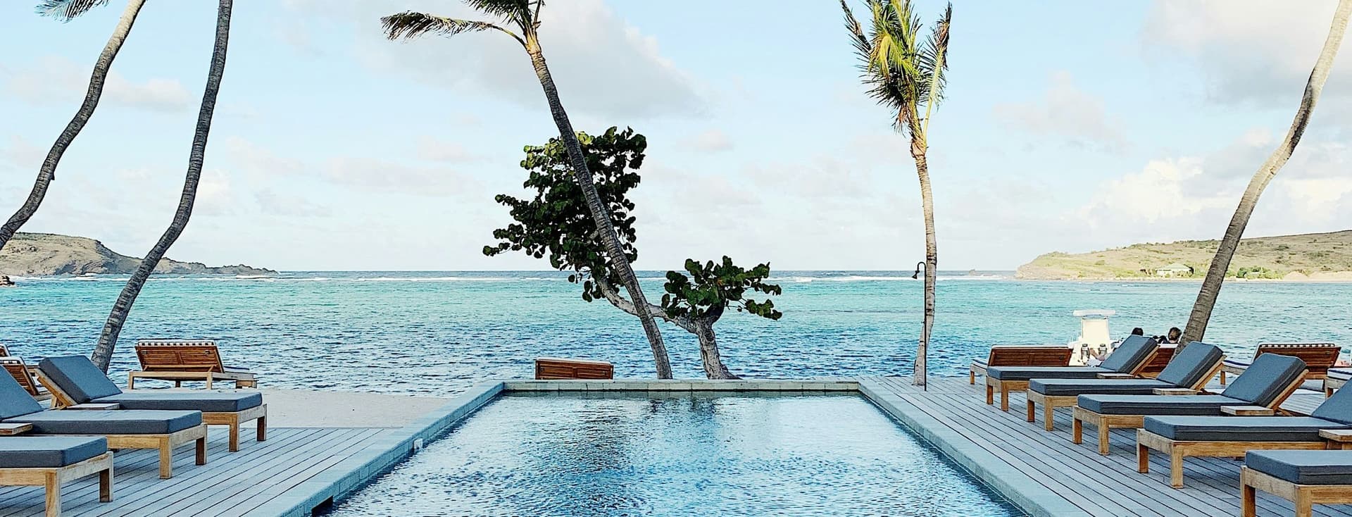 Infinity pool overlooking a turquoise ocean, flanked by palm trees and lounge chairs under a clear blue sky.