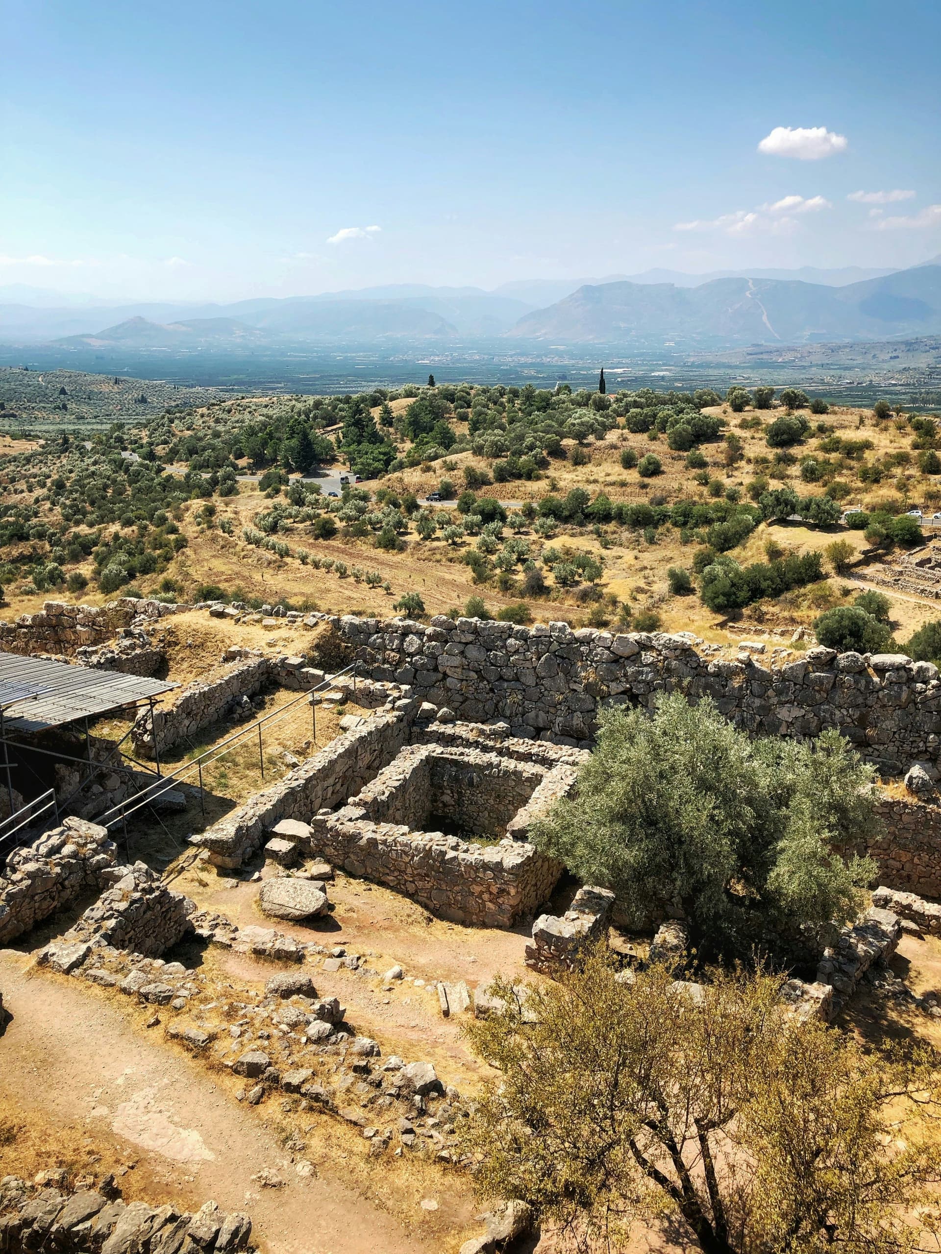 Ancient ruins of Mycenae, Greece, with stone walls, olive trees, and a distant view of mountains under a blue sky.