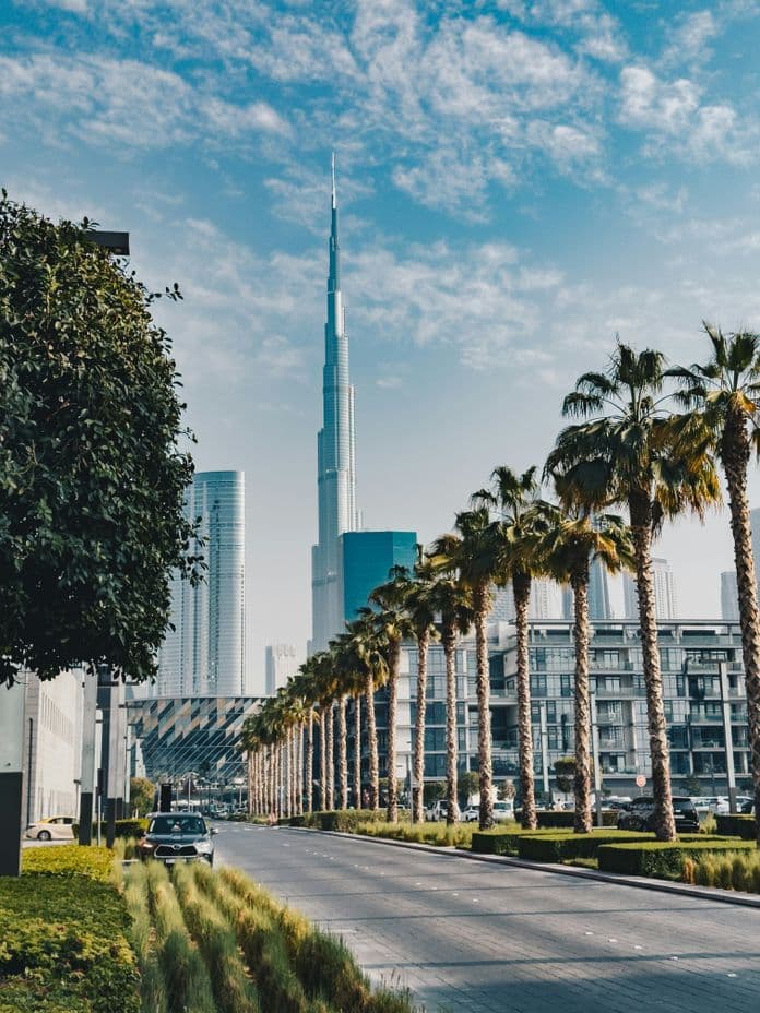 A cityscape featuring a tall skyscraper, palm-lined street, modern buildings, and a blue sky with scattered clouds.