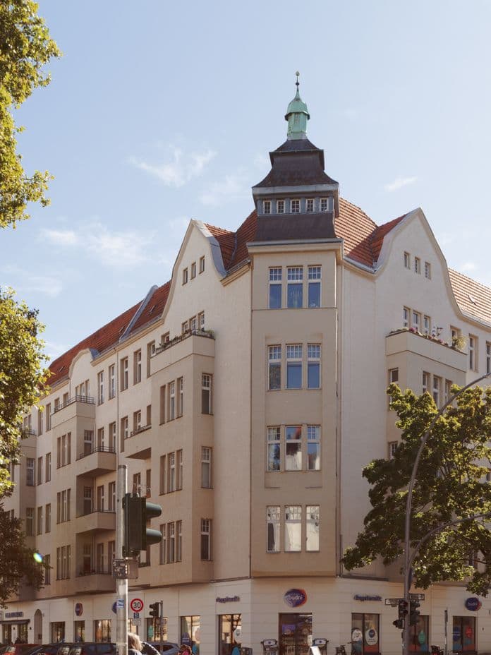Historic multi-story building with a corner tower, red-tiled roof, and shops at street level, surrounded by trees under a clear blue sky.