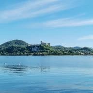Lake Maggiore with calm waters reflecting the blue sky. In the background, the Rocca di Angera stands atop a green hill, surrounded by mountainous and wooded landscapes.