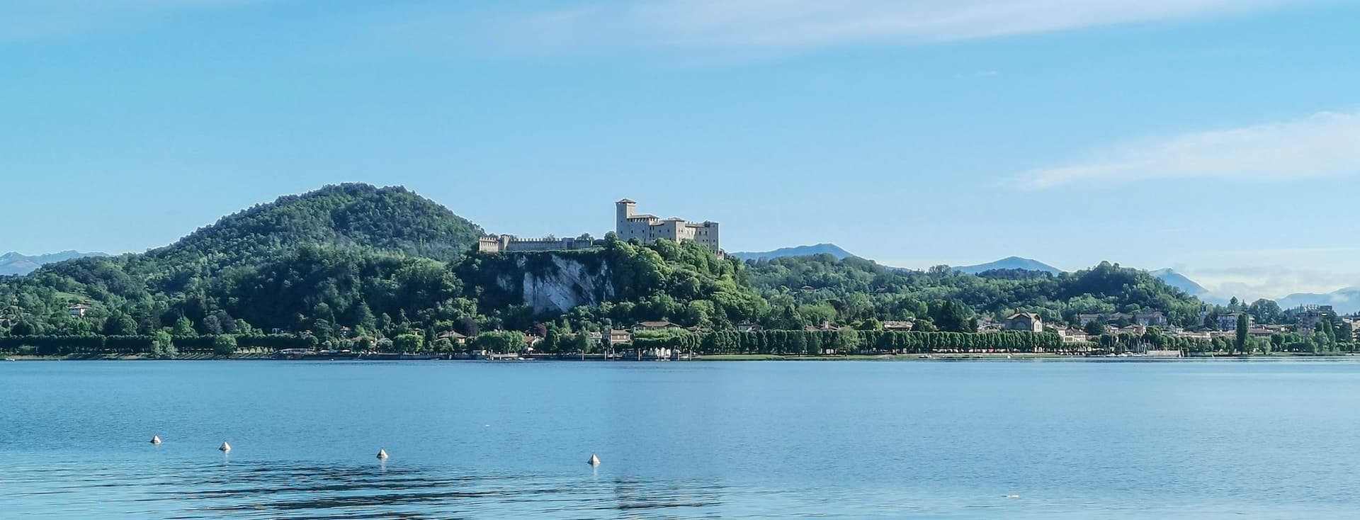 Lake Maggiore with calm waters reflecting the blue sky. In the background, the Rocca di Angera stands atop a green hill, surrounded by mountainous and wooded landscapes.