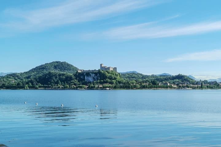 Il Lago Maggiore con acque calme che riflettono il cielo azzurro. Sullo sfondo, la Rocca di Angera si erge su una collina verde, circondata da paesaggi montuosi e boschivi.
