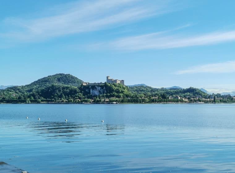 Der Lago Maggiore mit seinem ruhigen Wasser, in dem sich der blaue Himmel spiegelt. Im Hintergrund erhebt sich die Rocca di Angera auf einem grünen Hügel, umgeben von einer bergigen und bewaldeten Landschaft.