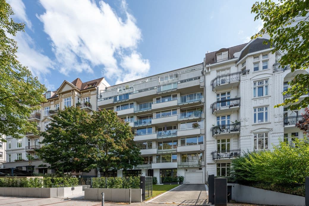 Street view of modern and traditional apartment buildings with balconies, surrounded by trees and greenery under a partly cloudy sky.