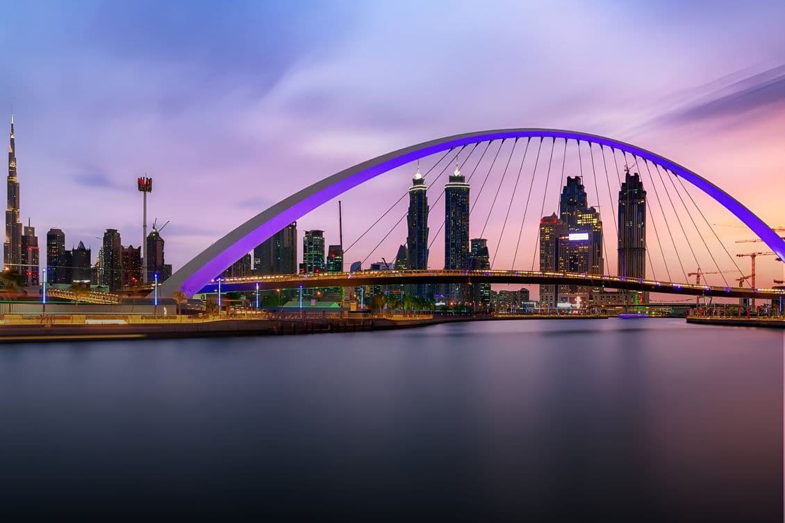 A city skyline at dusk with a prominent arched bridge over calm water, illuminated by colorful lights, and tall buildings in the background.