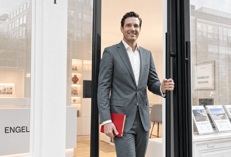 A friendly male real estate agent in a grey suit stands at the open glass door of an Engel & Völkers office, holding a red notebook and welcoming clients.