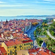 Aerial view of Nice, France, featuring red-tiled roofs, a Ferris wheel, and the Mediterranean Sea under a blue sky.