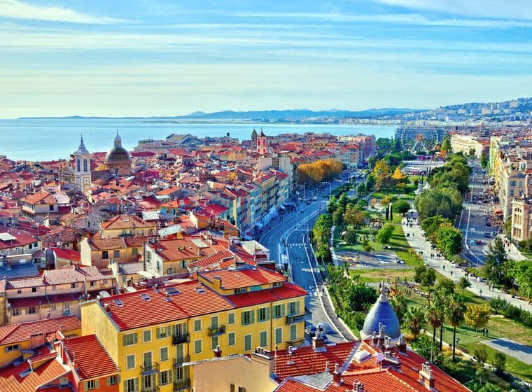 Aerial view of Nice, France, featuring red-tiled roofs, a Ferris wheel, and the Mediterranean Sea under a blue sky.