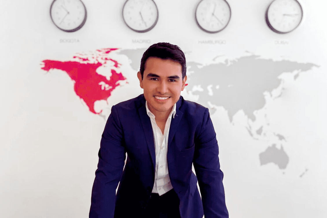 Smiling real estate agent in a blue suit, standing in front of a world map with clocks showing times in Bogota, Madrid, Hamburg, and Dubai.
