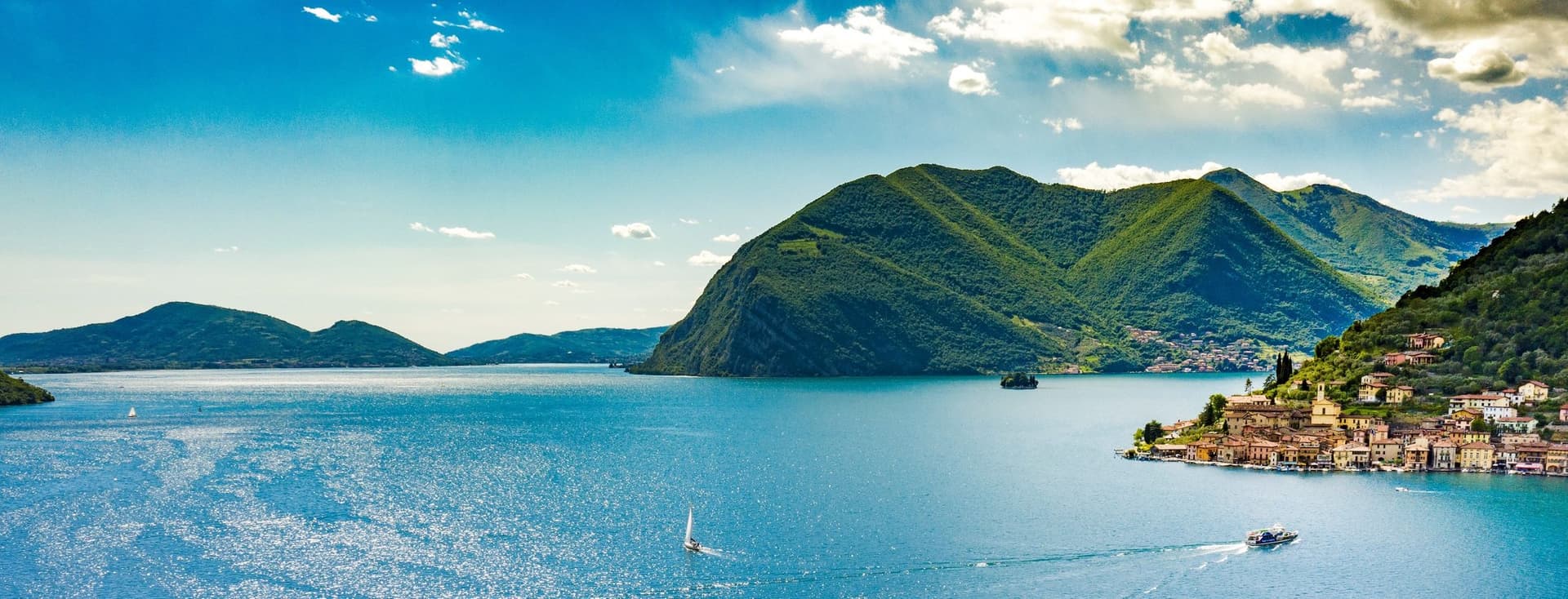 Malerischer Blick auf einen See mit Segelbooten, umgeben von üppig grünen Bergen und einem kleinen Dorf auf der rechten Seite unter einem klaren blauen Himmel.