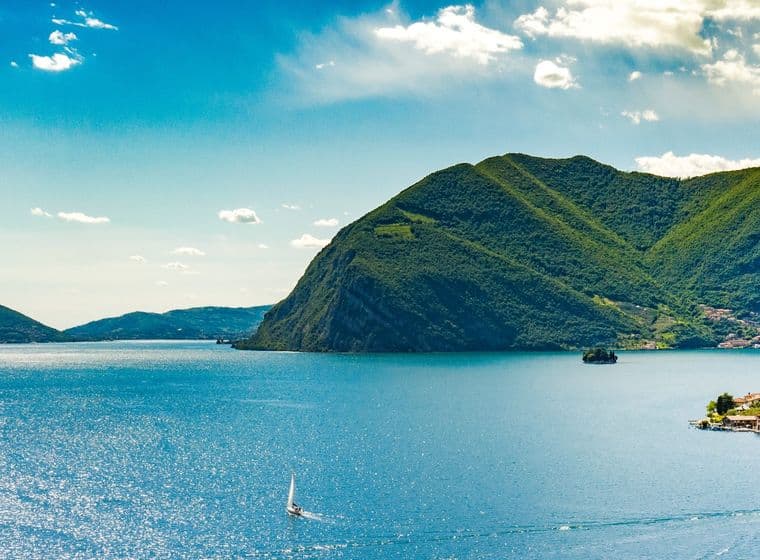 Malerischer Blick auf einen See mit Segelbooten, umgeben von üppig grünen Bergen und einem kleinen Dorf auf der rechten Seite unter einem klaren blauen Himmel.