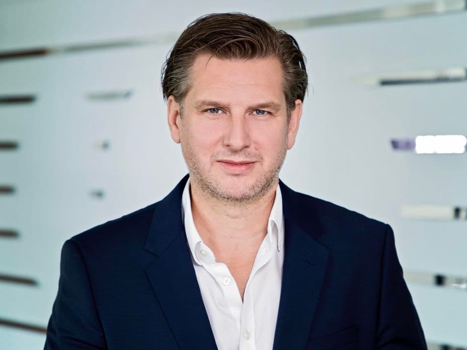Man in a navy suit with a white shirt, standing against a modern white background, looking at the camera with a neutral expression.