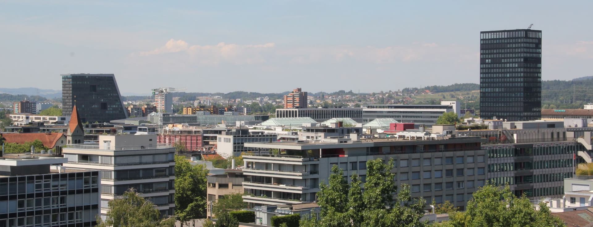 Skyline-Blick auf eine Stadt mit modernen Gebäuden, einem hohen schwarzen Turm und üppigem Grün unter klarem blauen Himmel.
