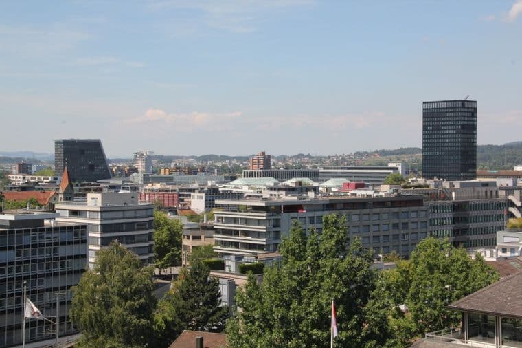 Vista dello skyline di una città con edifici moderni, un'alta torre nera e una vegetazione lussureggiante sotto un cielo azzurro e terso.