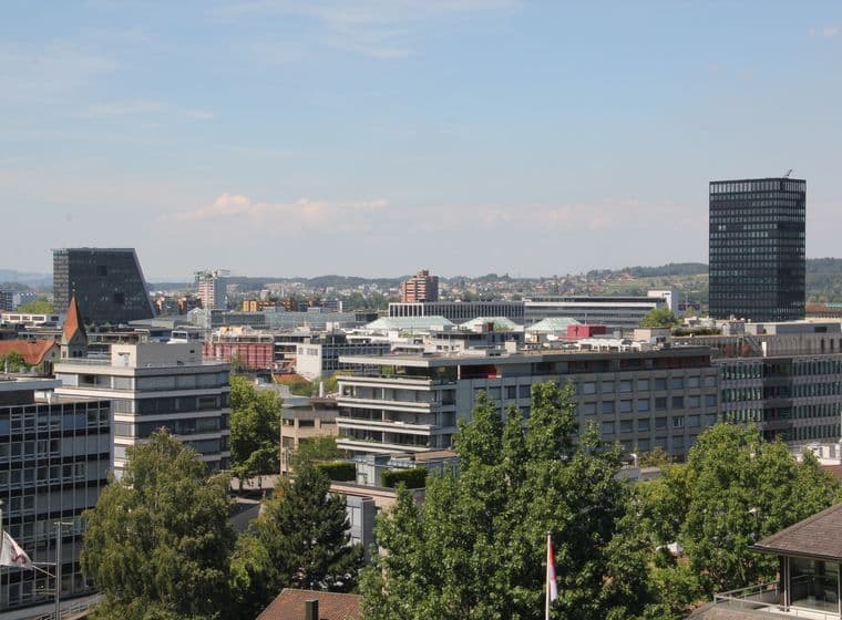 Vue aérienne d'une ville avec des bâtiments modernes, une haute tour noire et une végétation luxuriante sous un ciel bleu clair.
