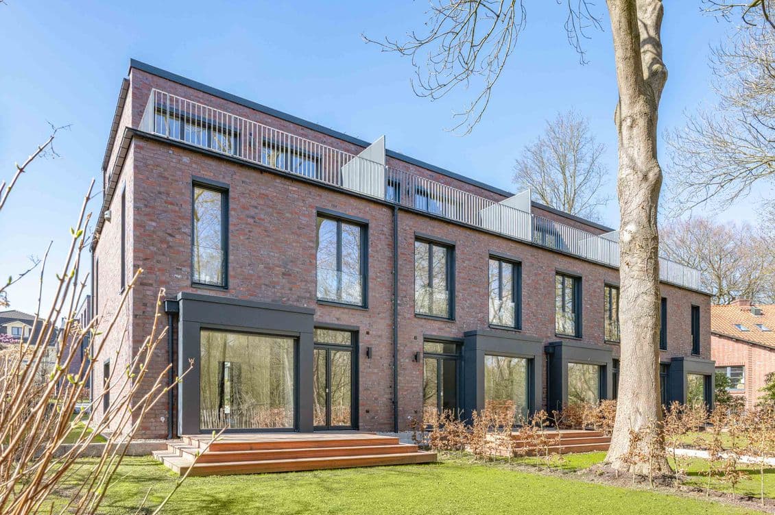 Modernes Backstein-Stadthaus mit großen Fenstern, umgeben von Bäumen und Grün unter klarem blauen Himmel.
