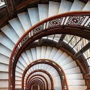 Spiral staircase with ornate railings, viewed from below, creating a tunnel-like effect with intricate patterns and geometric symmetry.