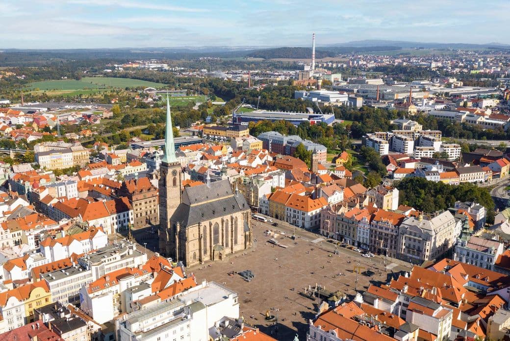 Aerial view of the historic center of Plzeň with Republic Square, dominated by St. Bartholomew’s Cathedral and its tall spire. Surrounding buildings have orange rooftops, with modern cityscape and greenery in the background.