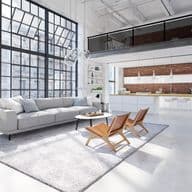 Bright, modern loft apartment with gray sofa, leather chairs, and large windows. A kitchen with brick wall is in the background.