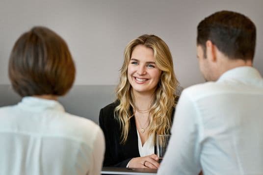 A smiling young woman with blonde wavy hair in professional attire sits across from two interviewers in white shirts during what appears to be a job interview in a modern office setting.