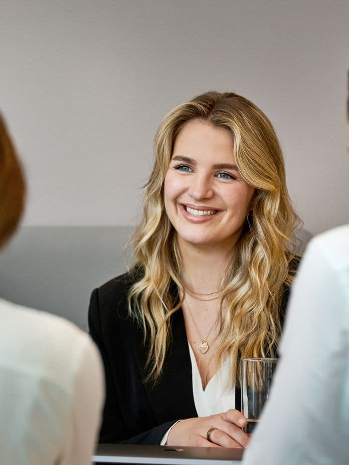 A smiling young woman with blonde wavy hair in professional attire sits across from two interviewers in white shirts during what appears to be a job interview in a modern office setting.