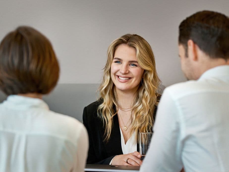 A smiling young woman with blonde wavy hair in professional attire sits across from two interviewers in white shirts during what appears to be a job interview in a modern office setting.
