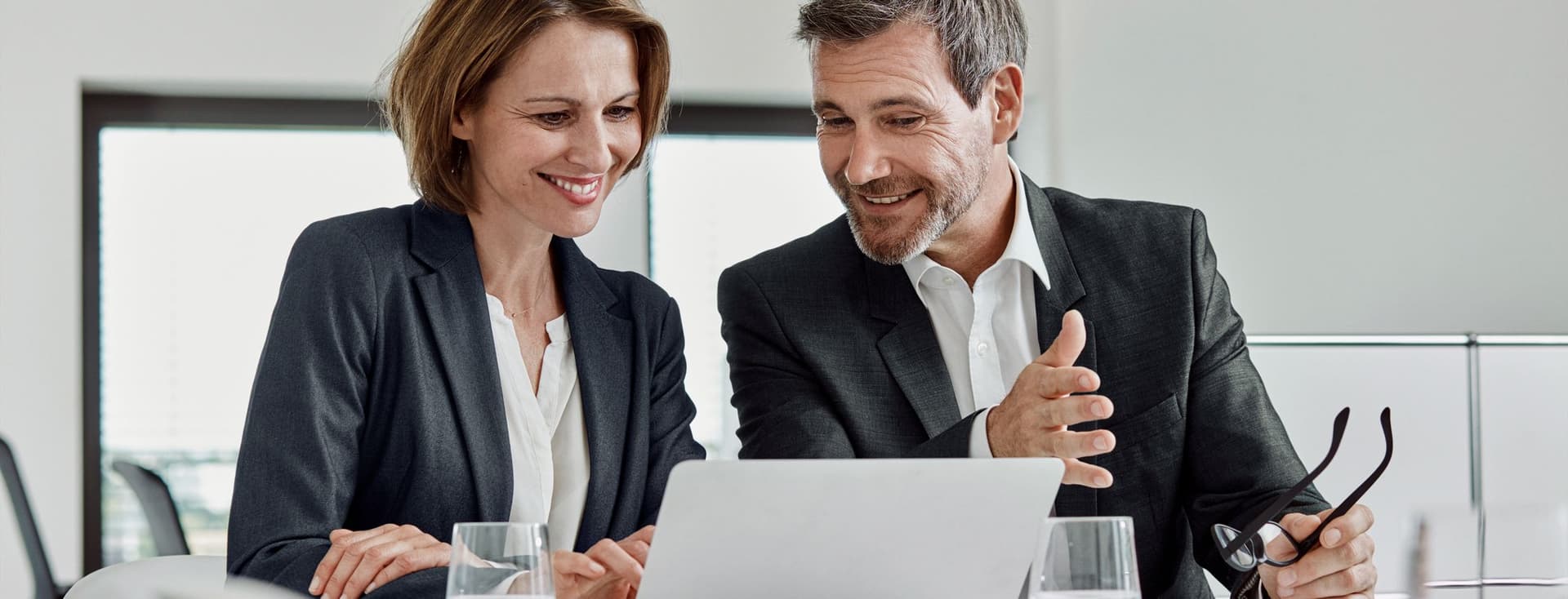 Un home i una dona somrients amb vestits miren un portàtil sobre una taula blanca en una oficina lluminosa.