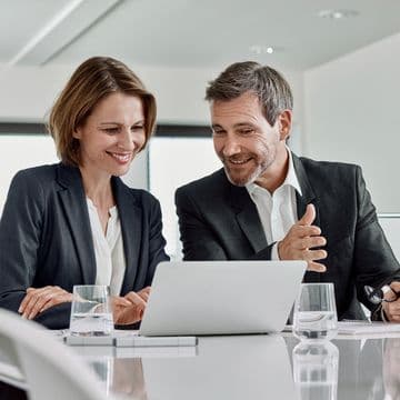 Two business professionals, a woman and a man both wearing dark blazers with white shirts, are smiling while looking at a laptop screen in a modern office setting. The scene captures a moment of positive collaboration with glasses of water on the table and natural lighting in the background.
