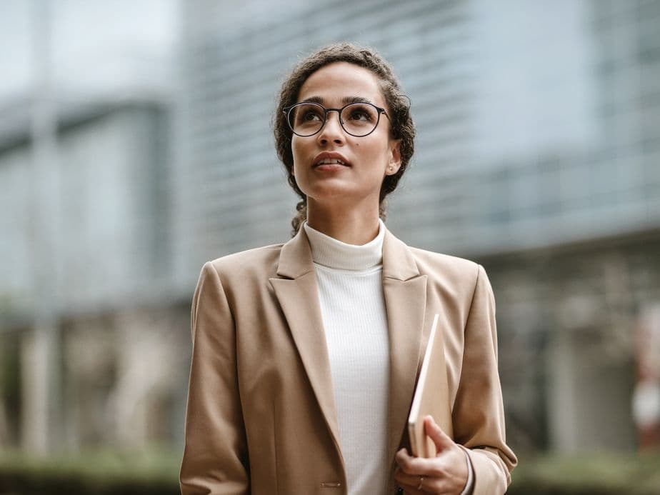 A confident businesswoman wearing glasses, a beige blazer, and white turtleneck stands in front of a modern building. She holds documents while looking upward with an optimistic expression, her hair styled in a professional updo.