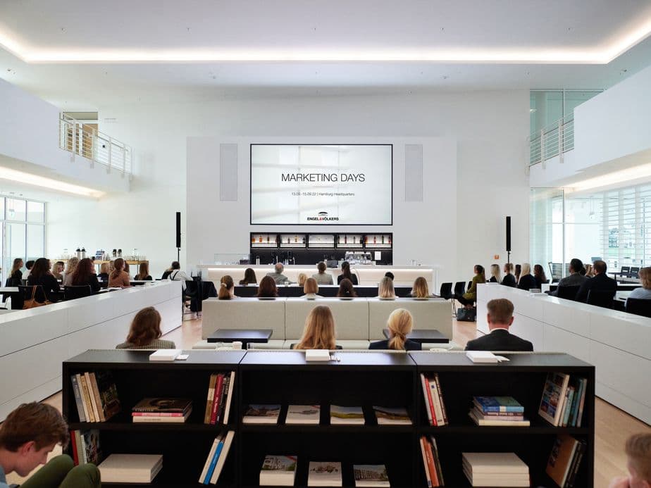 View of a modern white conference room with tiered seating filled with attendees watching a presentation screen displaying 'Marketing Days', featuring built-in bookshelves and modern lighting