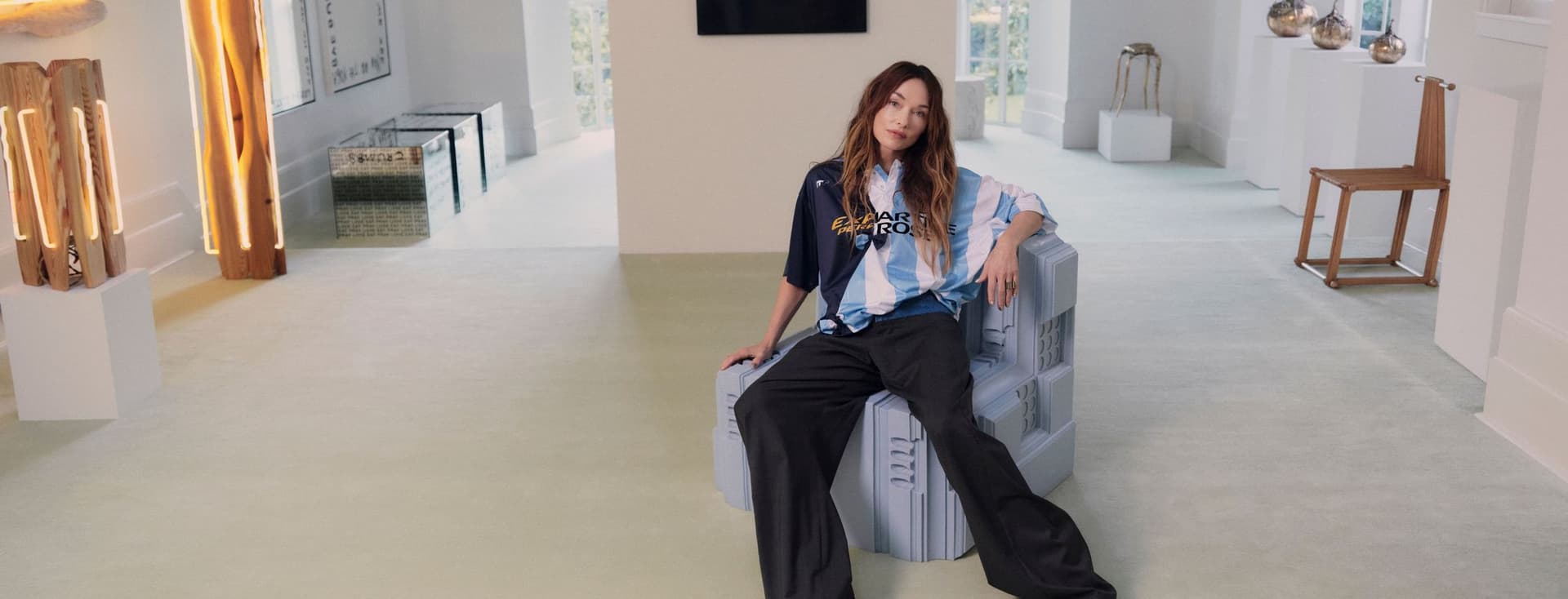 Eine stilvolle Frau in einem blau gestreiften Hemd und einer schwarzen Hose sitzt auf einer geometrischen Bank in einem modernen, mit Kunstwerken gefüllten Raum.