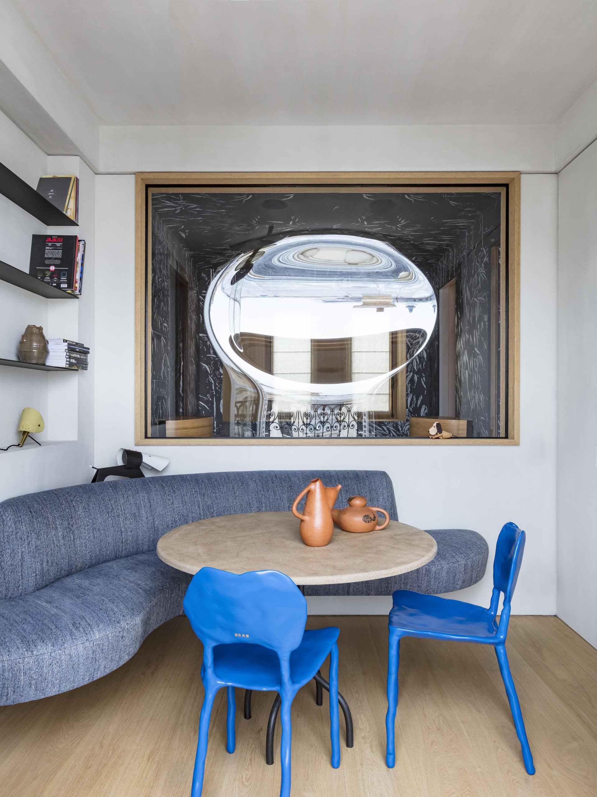Un coin repas avec un canapé incurvé bleu, une table ronde et des chaises bleues. Un grand luminaire réfléchissant est suspendu au-dessus, reflétant la pièce.