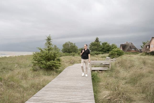 Mann, der auf einem Holzpfad durch grasbewachsene Dünen geht, mit bewölktem Himmel und Häusern im Hintergrund.