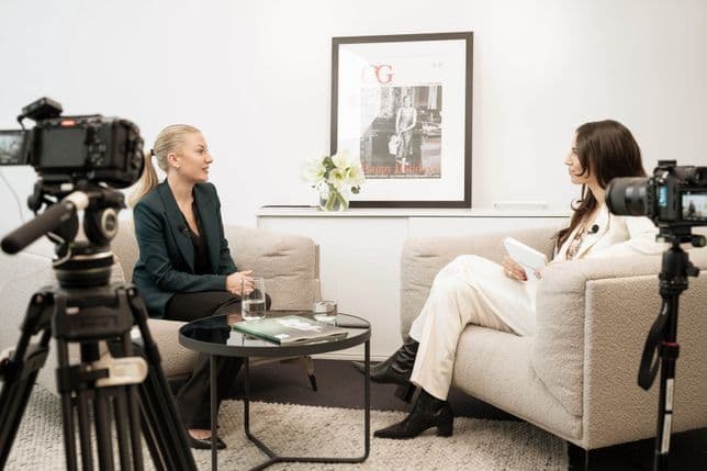 Two women are engaged in an interview in a modern setting with cameras around them, sitting on beige chairs near a table with water glasses.