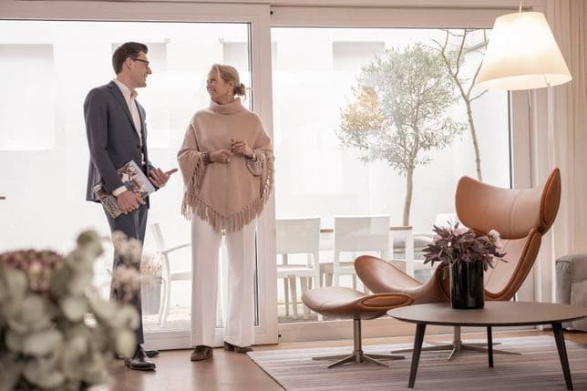 Ein Mann und eine Frau stehen in einem modernen Wohnzimmer und unterhalten sich. Sie befinden sich in der Nähe eines großen Fensters mit Blick auf eine Terrasse und einen Baum.