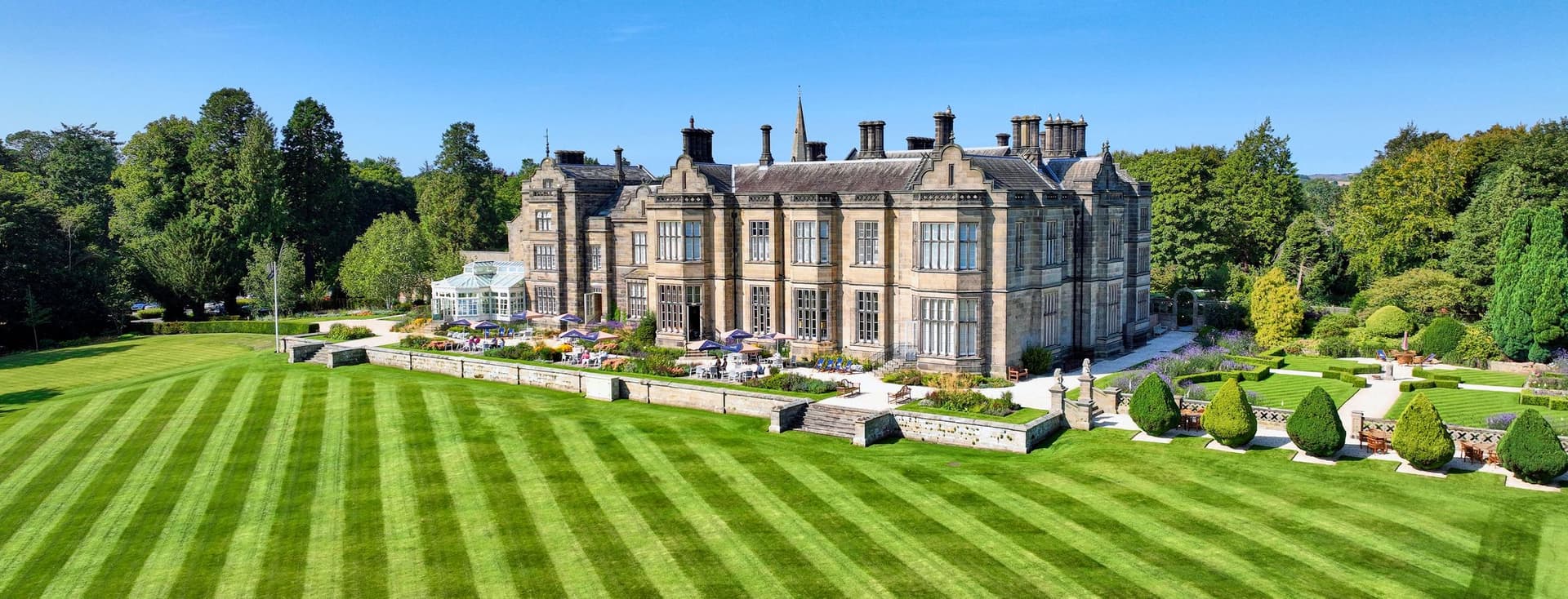 Aerial view of a large, stately stone manor house with manicured lawns and gardens under a clear blue sky.