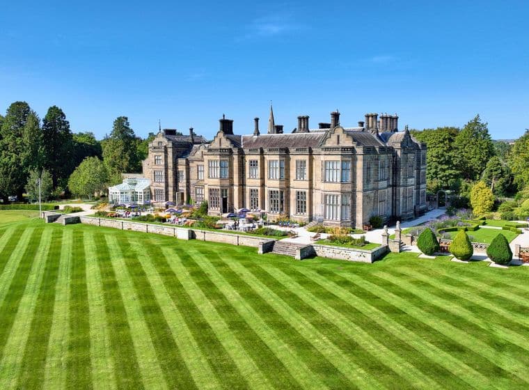 Aerial view of a large, stately stone manor house with manicured lawns and gardens under a clear blue sky.
