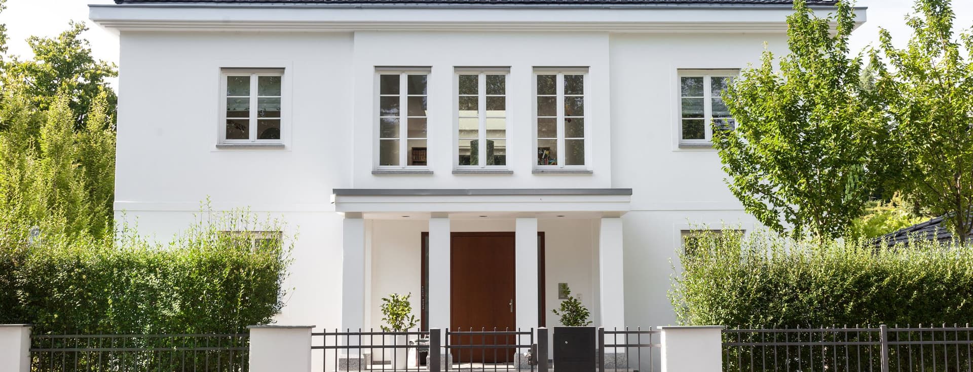 Two-story white city villa with hipped roof, large entrance door and well-maintained front garden behind a metal fence