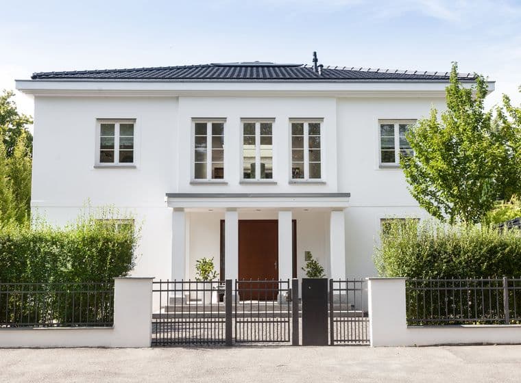 Two-story white city villa with hipped roof, large entrance door and well-maintained front garden behind a metal fence
