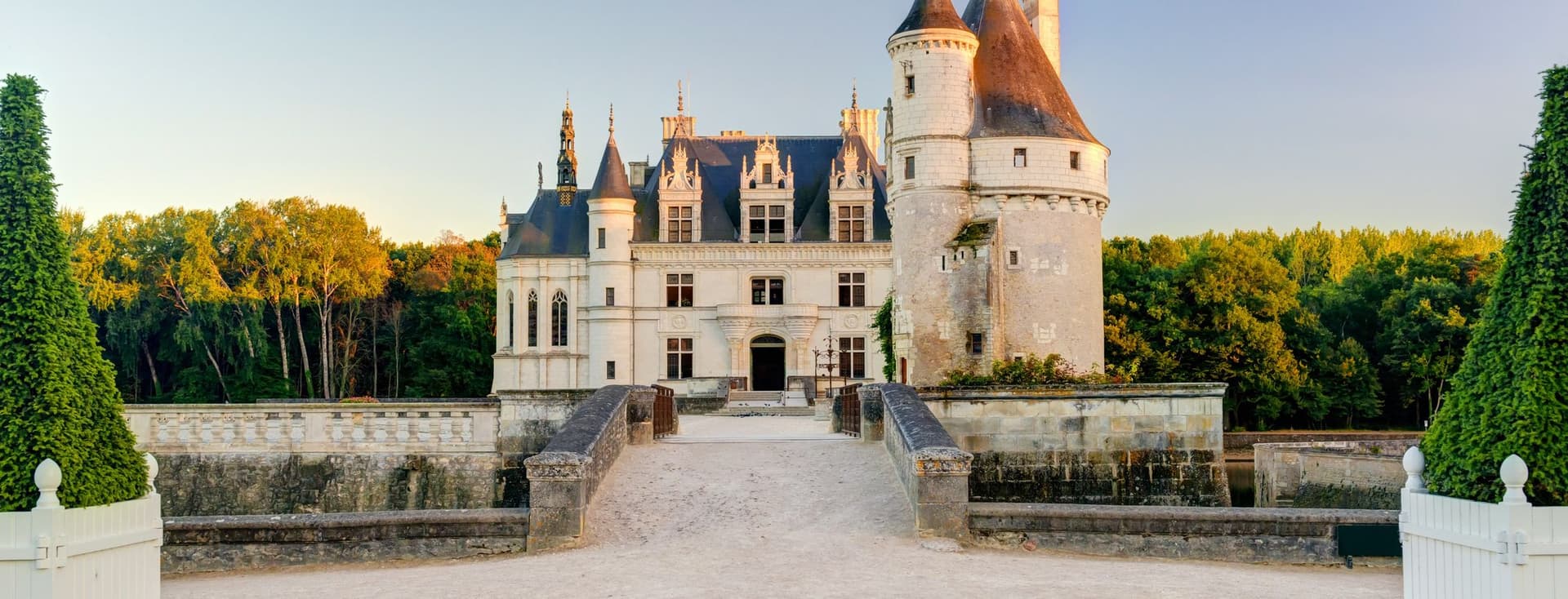 Château de Chenonceau, a white stone castle with turrets, towers, and a blue roof, surrounded by green trees under a clear blue sky.