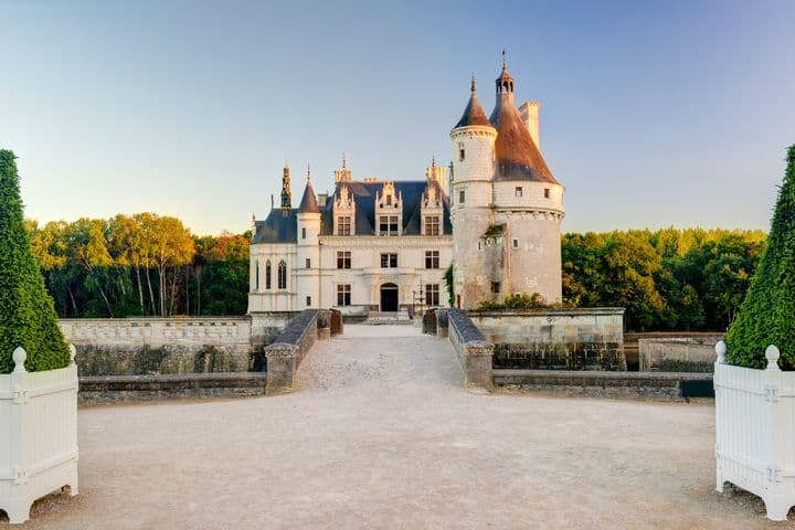 Château de Chenonceau, a white stone castle with turrets, towers, and a blue roof, surrounded by green trees under a clear blue sky.