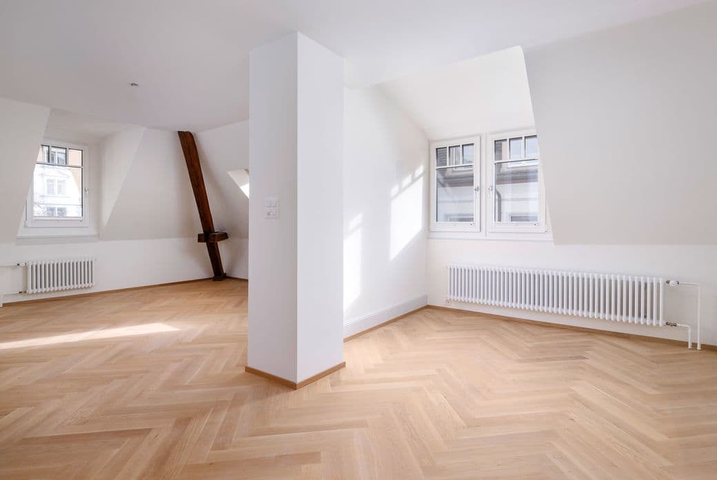 Bright, empty room with herringbone wood floors, white walls, and two windows. A white column stands in the center.